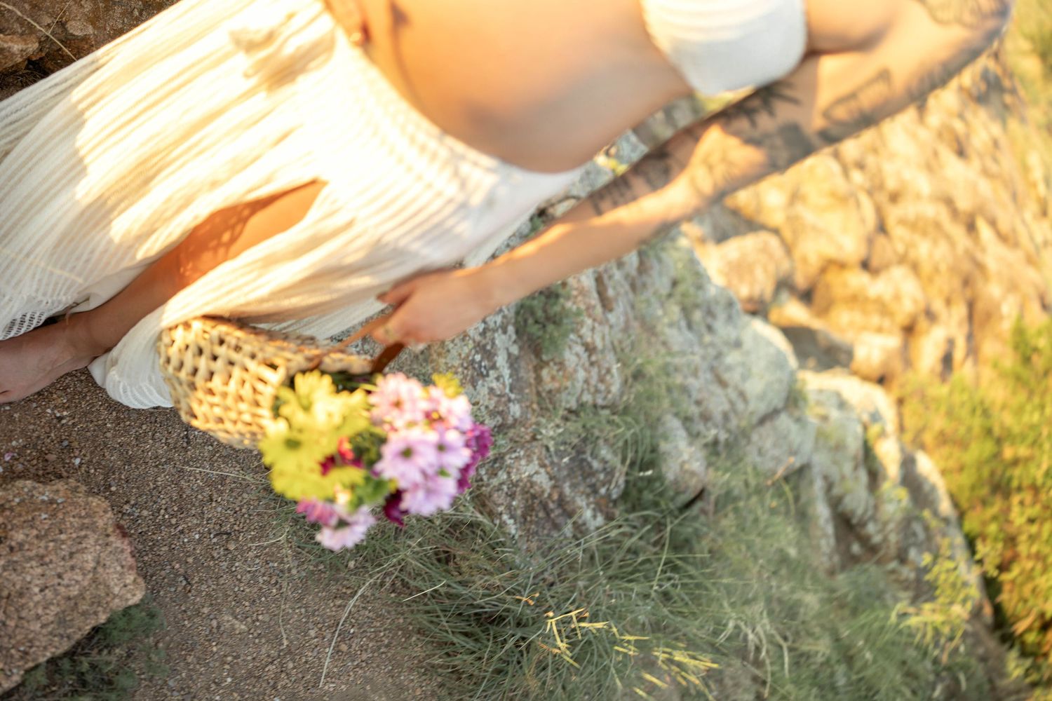 Woman in white dress kneeling on rocky terrain, holding a wicker bag with pink and yellow wildflowers in golden sunlight.