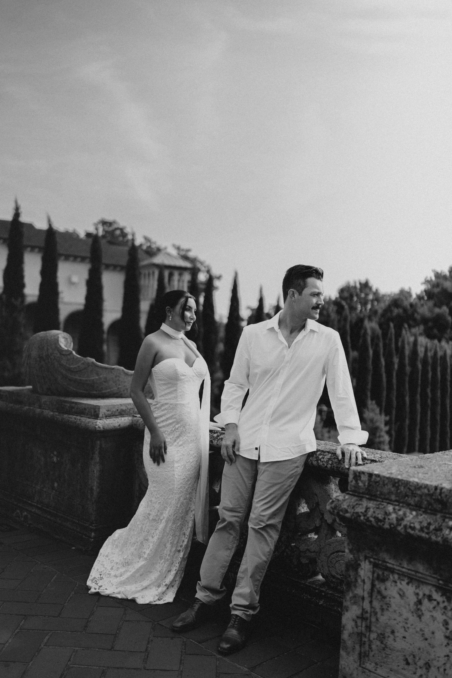 Black and white photo of a couple in wedding attire posing on a stone terrace with cypress trees backdrop.