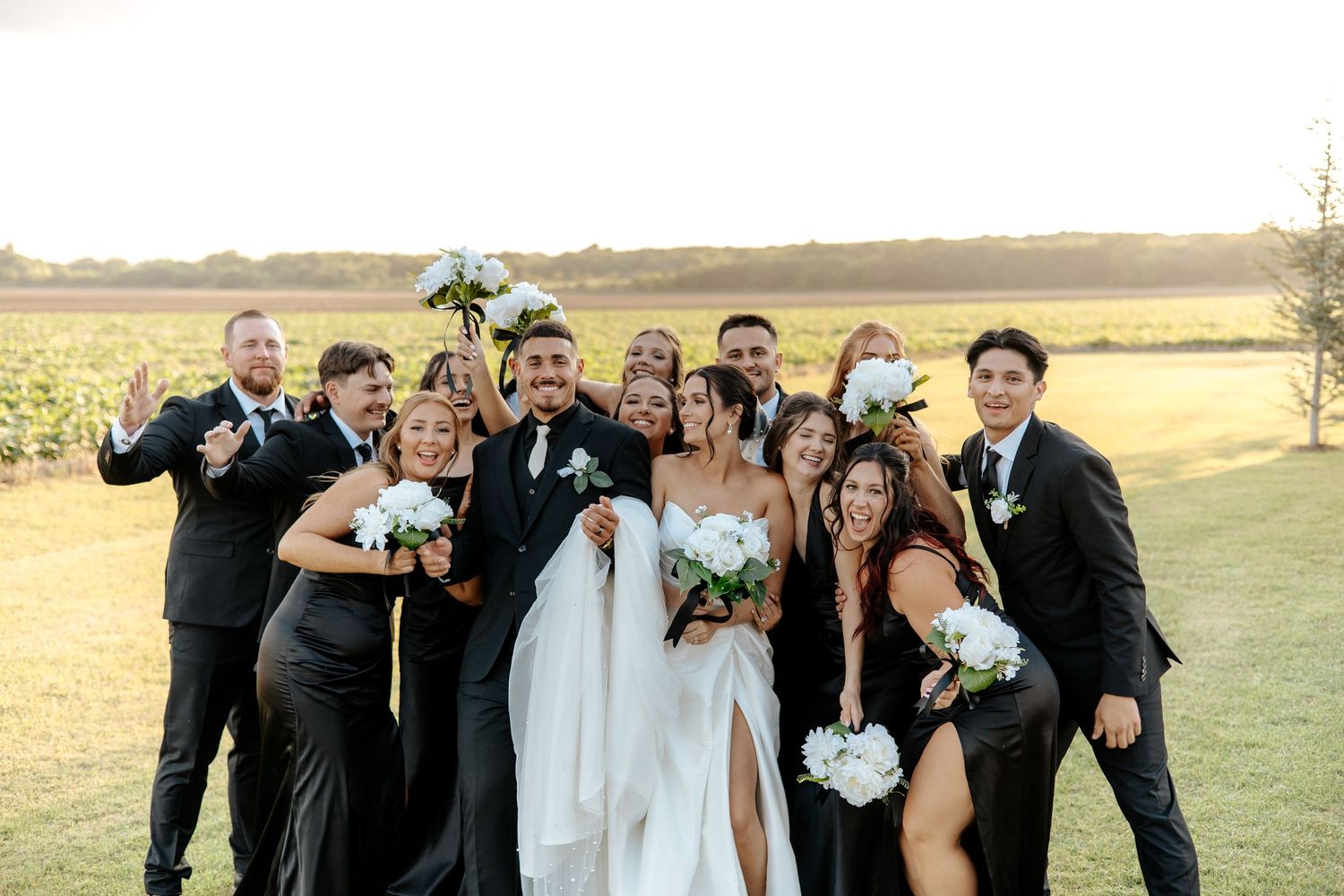 Bride and groom celebrate with wedding party in black attire at golden hour outdoor venue.