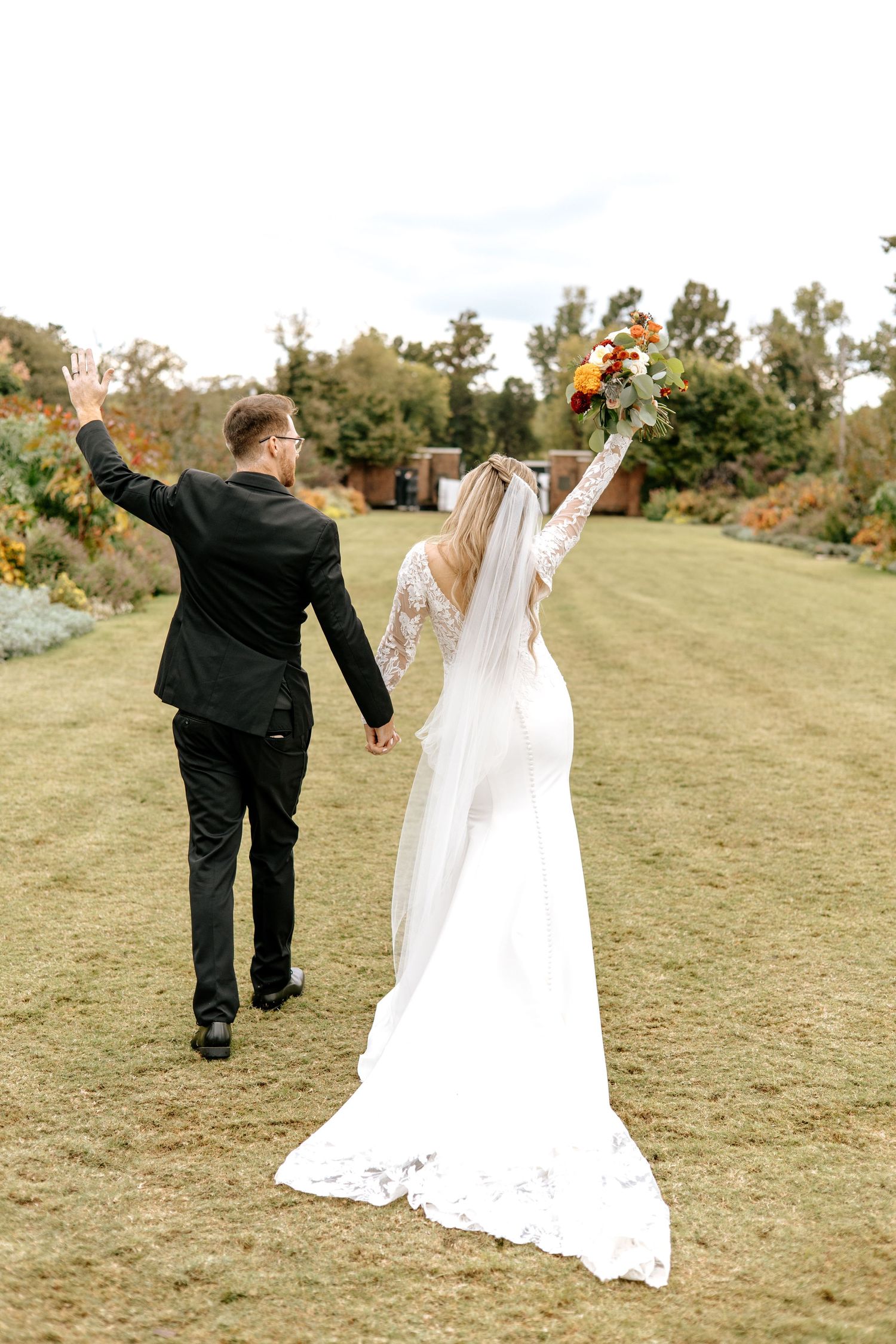 Bride and groom walk away celebrating with raised arms through colorful fall garden setting.