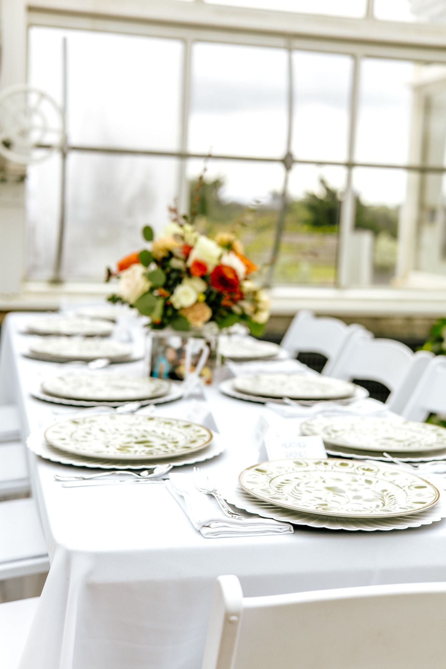 Elegant wedding reception table set with ornate plates, white linens, and colorful floral centerpiece in a greenhouse venue.