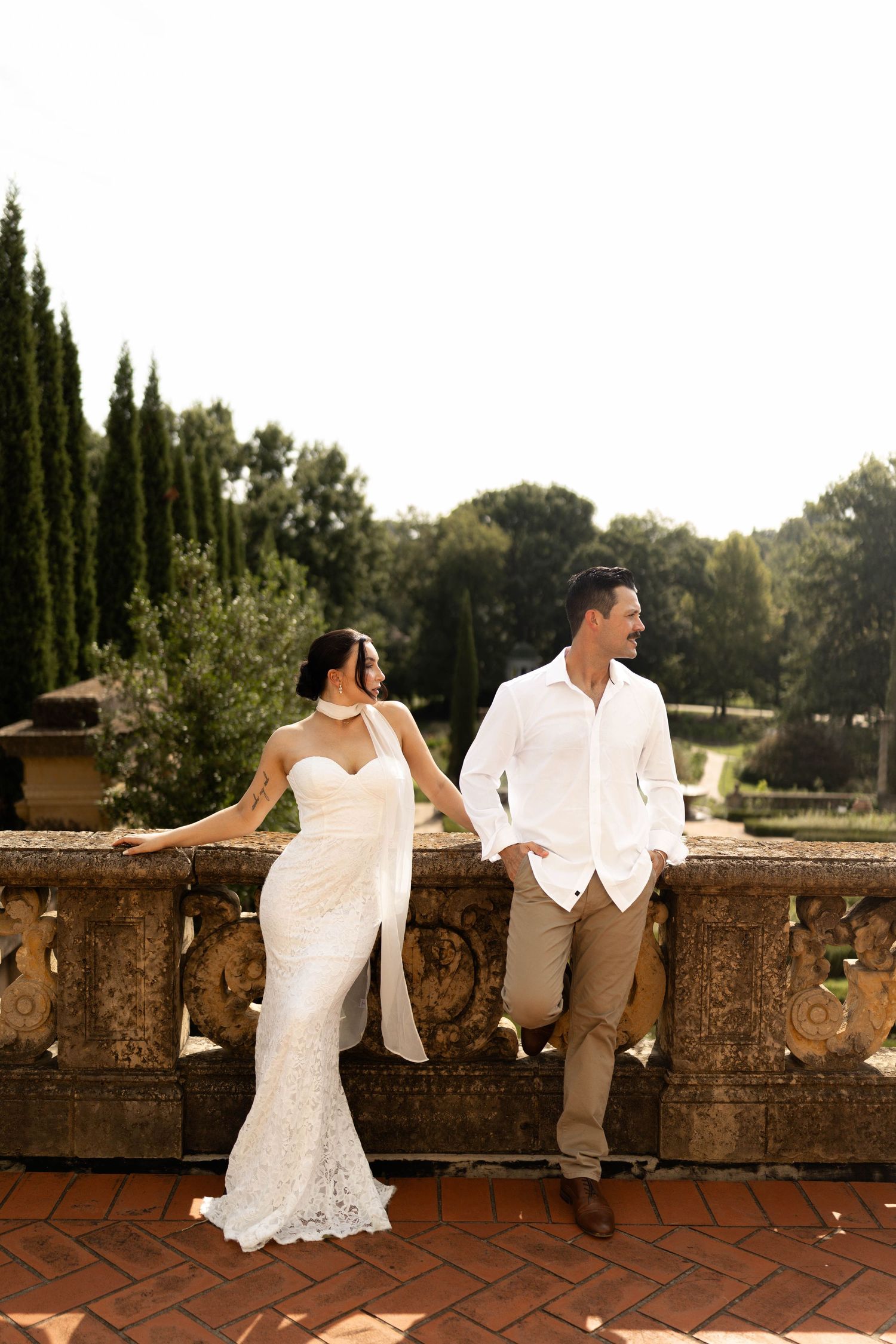 Bride in white lace gown and groom in white shirt hold hands on a stone balcony terrace in Tuscany.