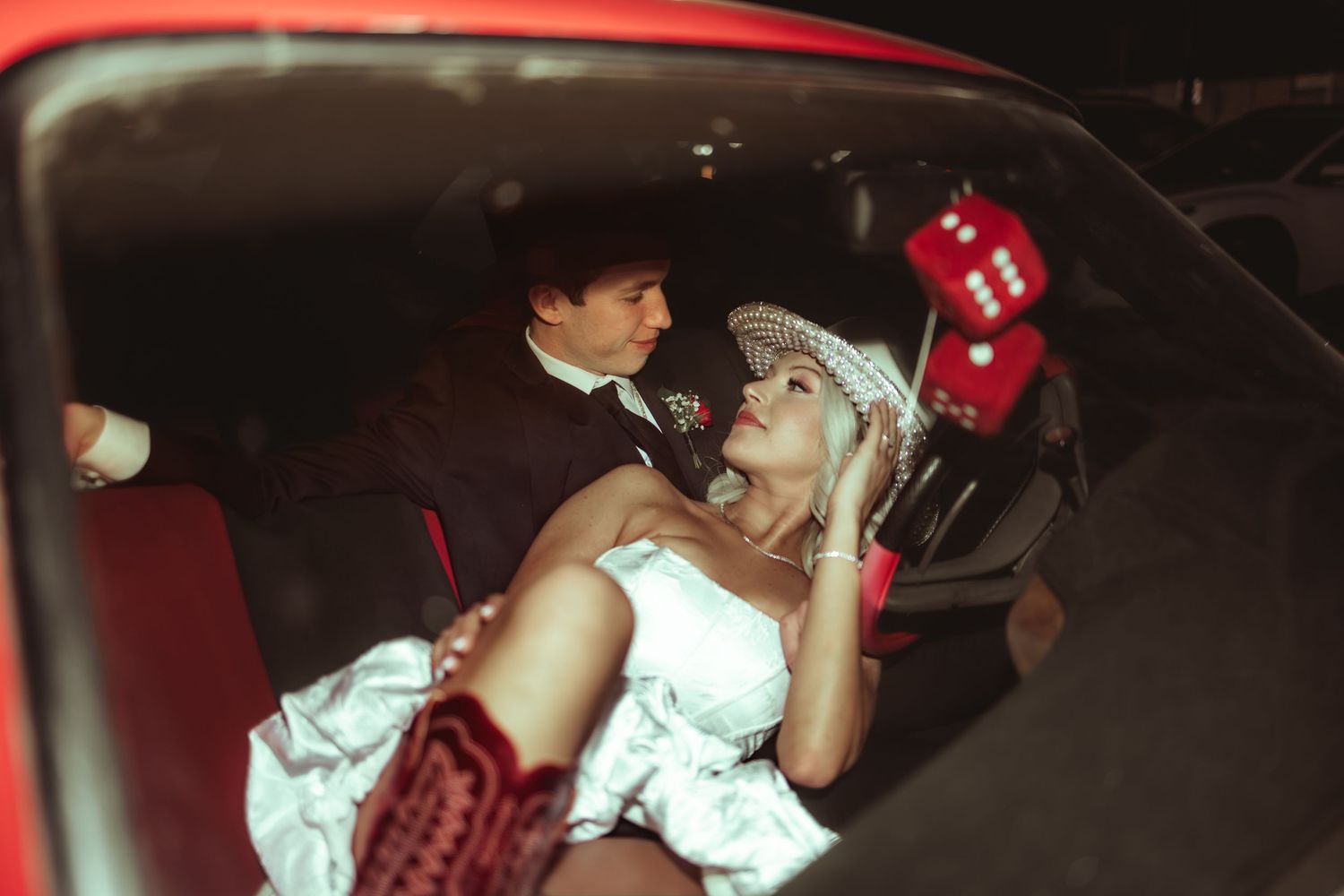 Bride and groom sitting in a vintage red car with fuzzy dice, sharing a romantic moment in wedding attire.