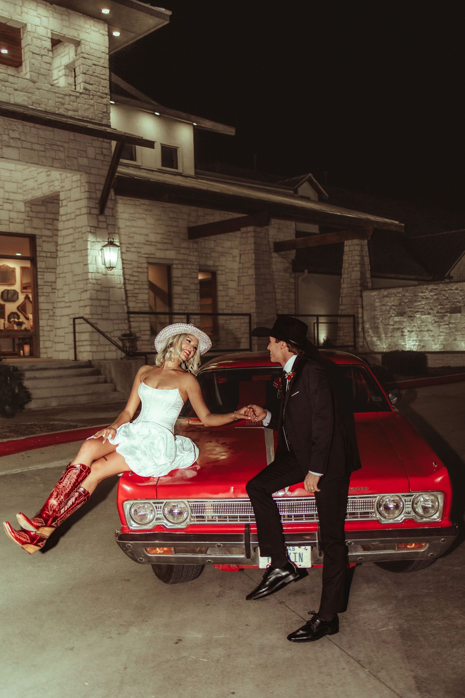 Couple in western attire poses on a red classic car at night outside a stone building, bride in white dress and cowboy boots.