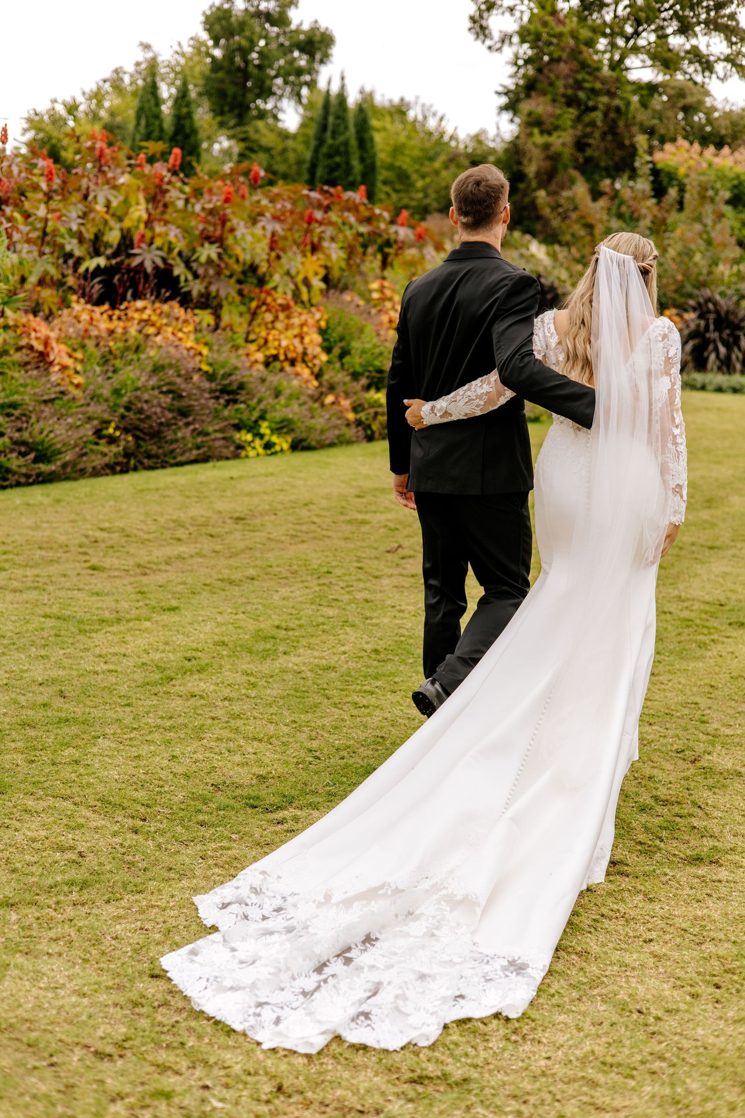 Bride and groom walk away through lush garden, bride wearing long lace-trimmed cathedral veil and fitted white gown.
