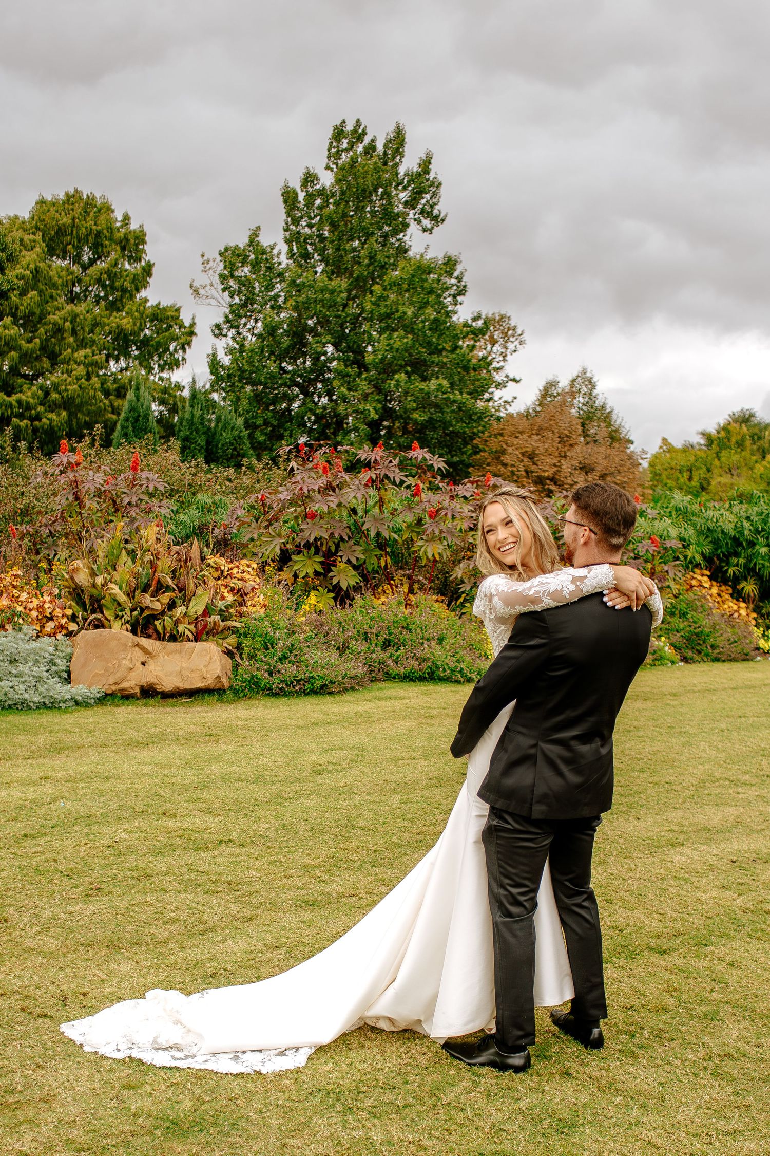 Bride in long-train lace wedding gown dances with groom amid autumn garden colorful foliage.