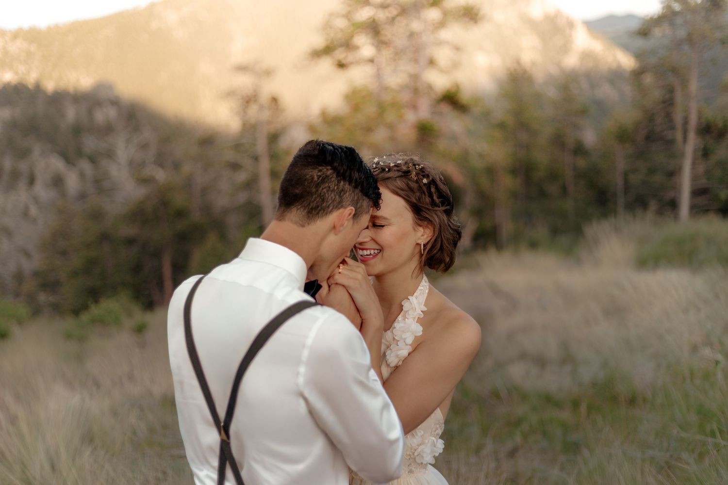 Romantic couple nearly kissing in sun-drenched meadow with mountains behind, groom in suspenders and bride in lace gown.