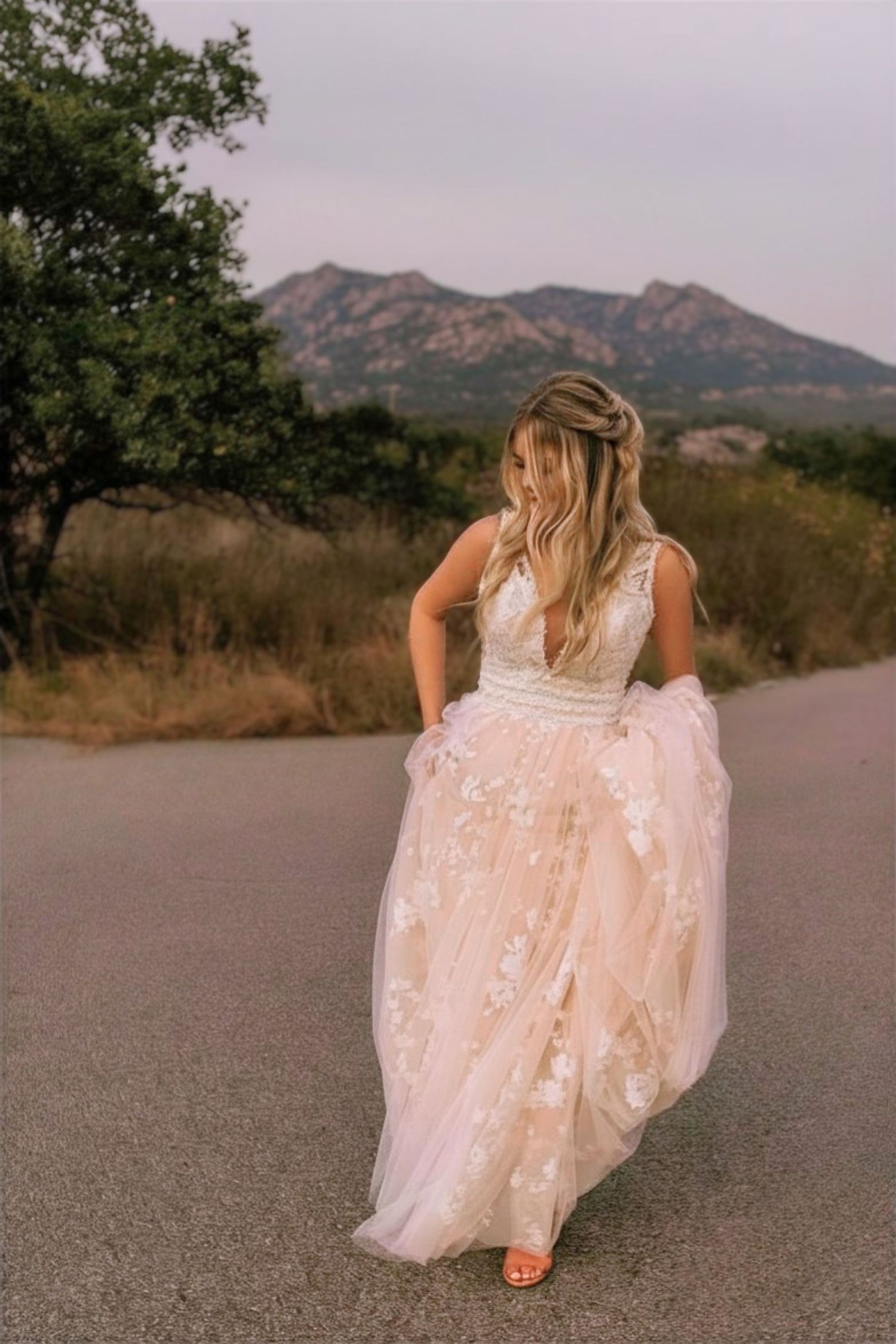 Bride in a flowy blush lace wedding gown walking on a scenic road with rocky mountains and trees in the background.