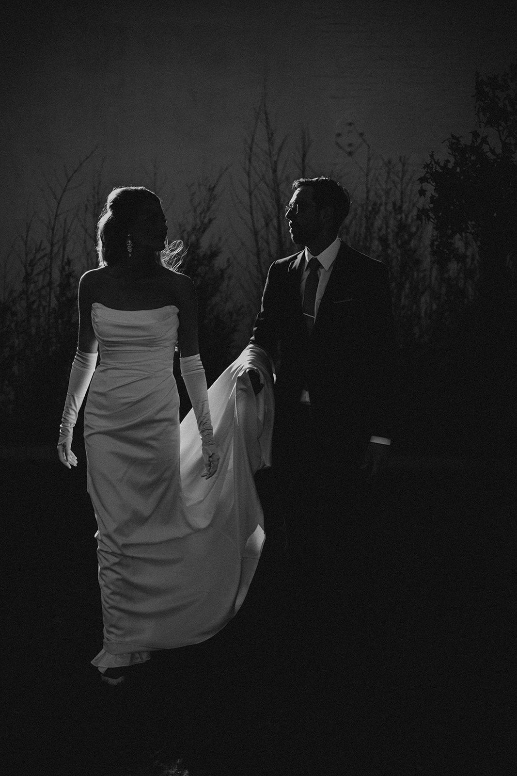 Dramatic black and white silhouette of bride and groom walking hand in hand at night.