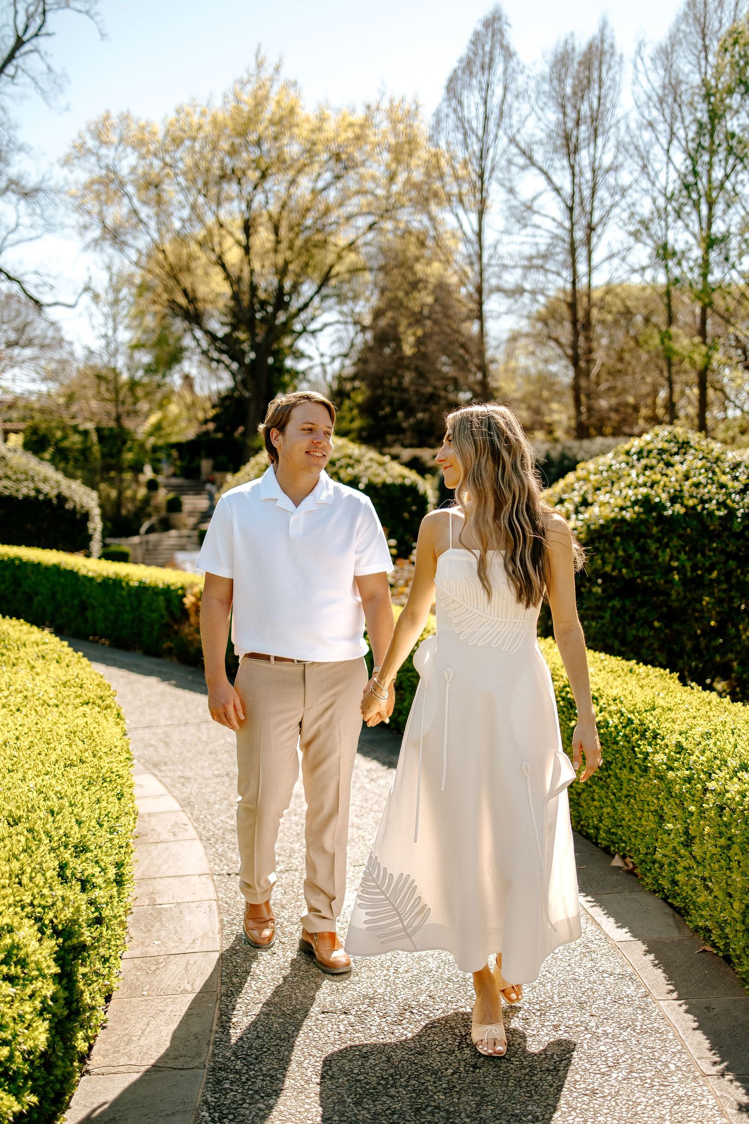 Couple holding hands walking through a sunny garden path, woman in white dress and man in white shirt and khaki pants.
