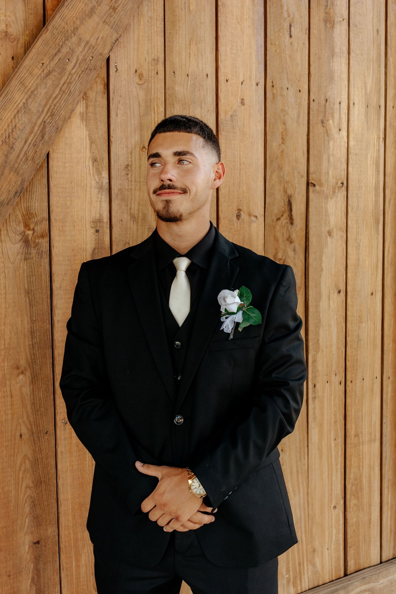 Groom in black suit with white tie and boutonniere poses against rustic wood wall.