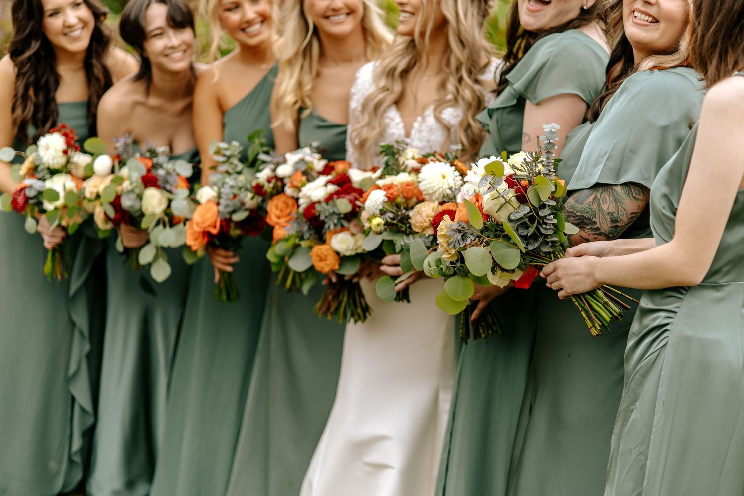 Bride surrounded by bridesmaids in sage green gowns, all holding lush autumn bouquets with orange, red, and white blooms.