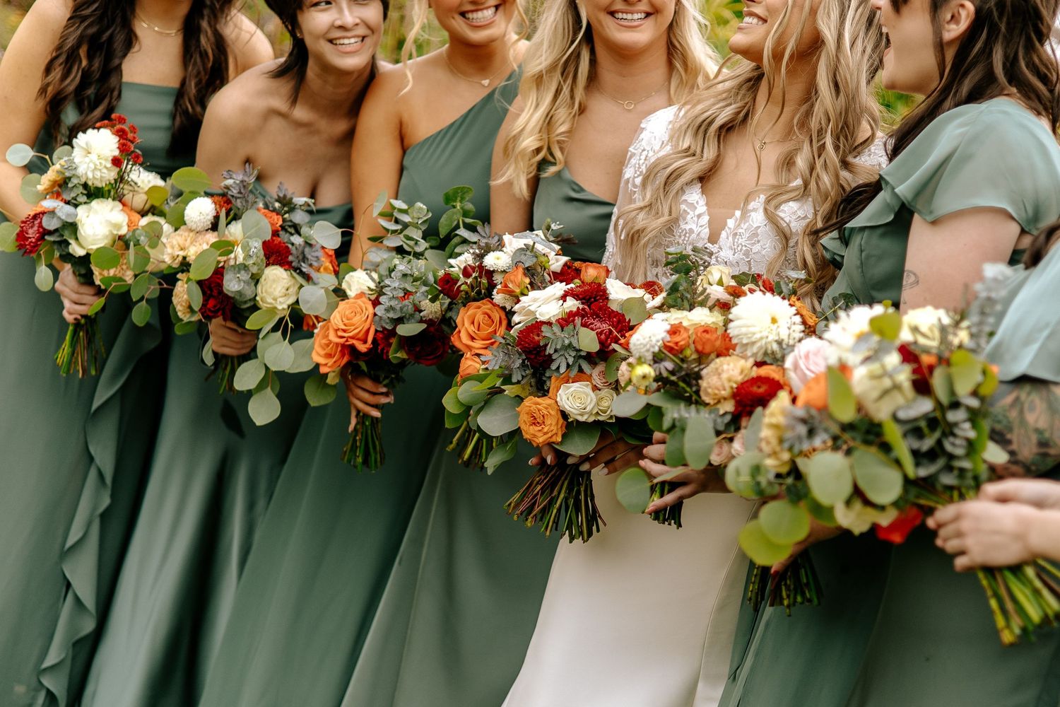 Bride with bridesmaids in sage green dresses holding colorful fall wedding bouquets outdoors.