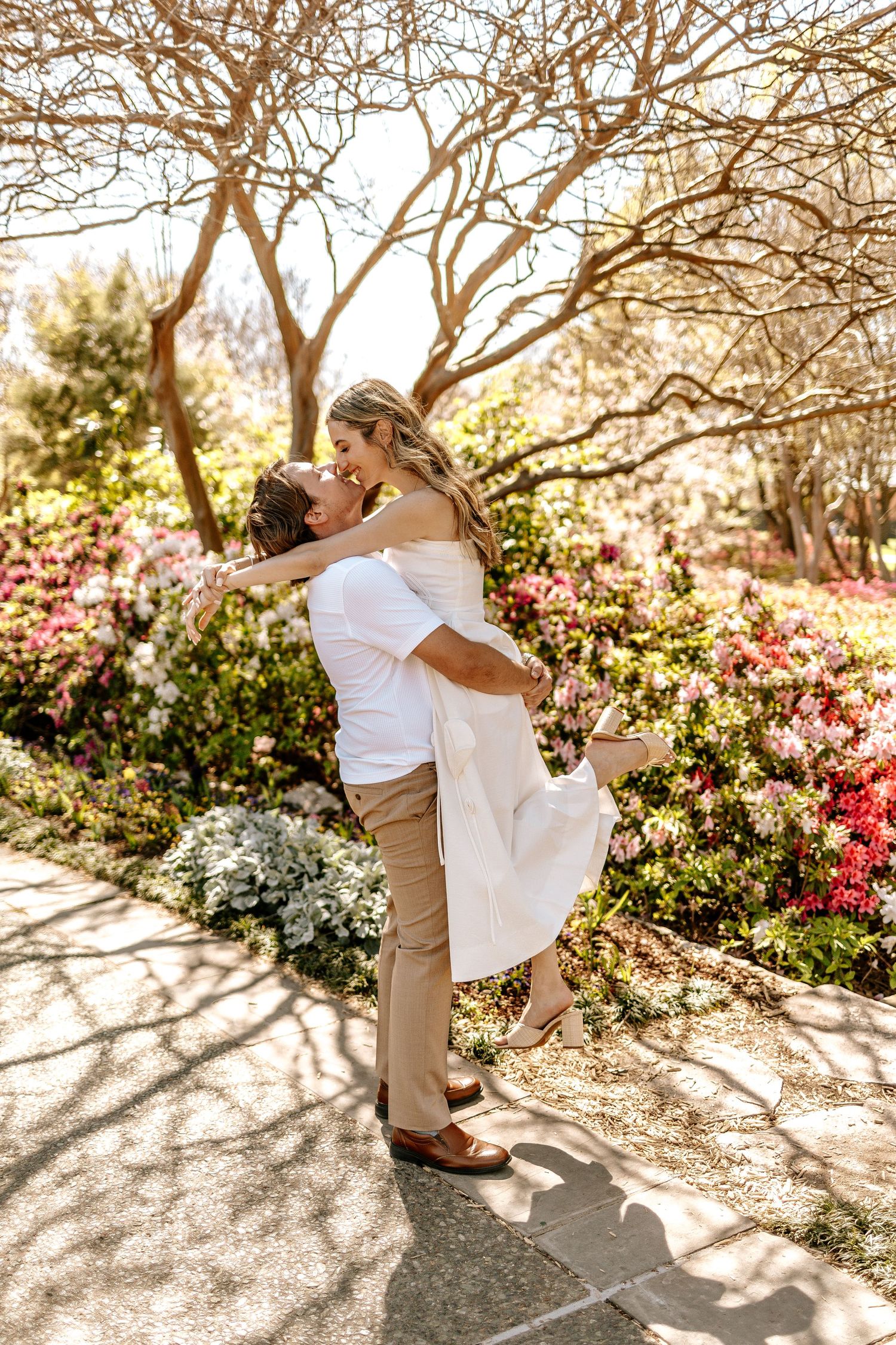 A couple shares a romantic kiss as the man lifts the woman in a colorful spring garden with blooming flowers.