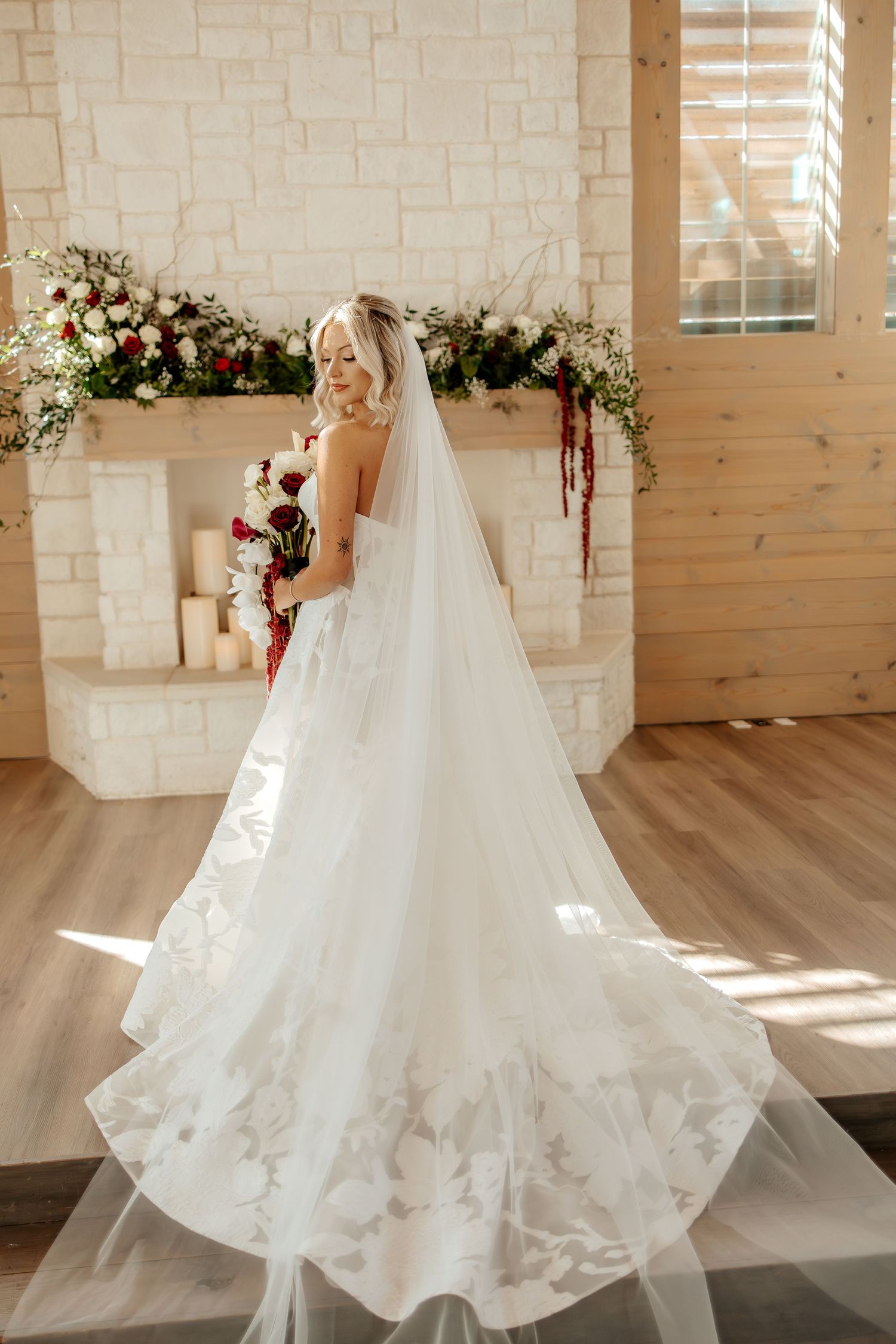 Blonde bride in a floral lace gown with a long cathedral veil standing before a stone fireplace.