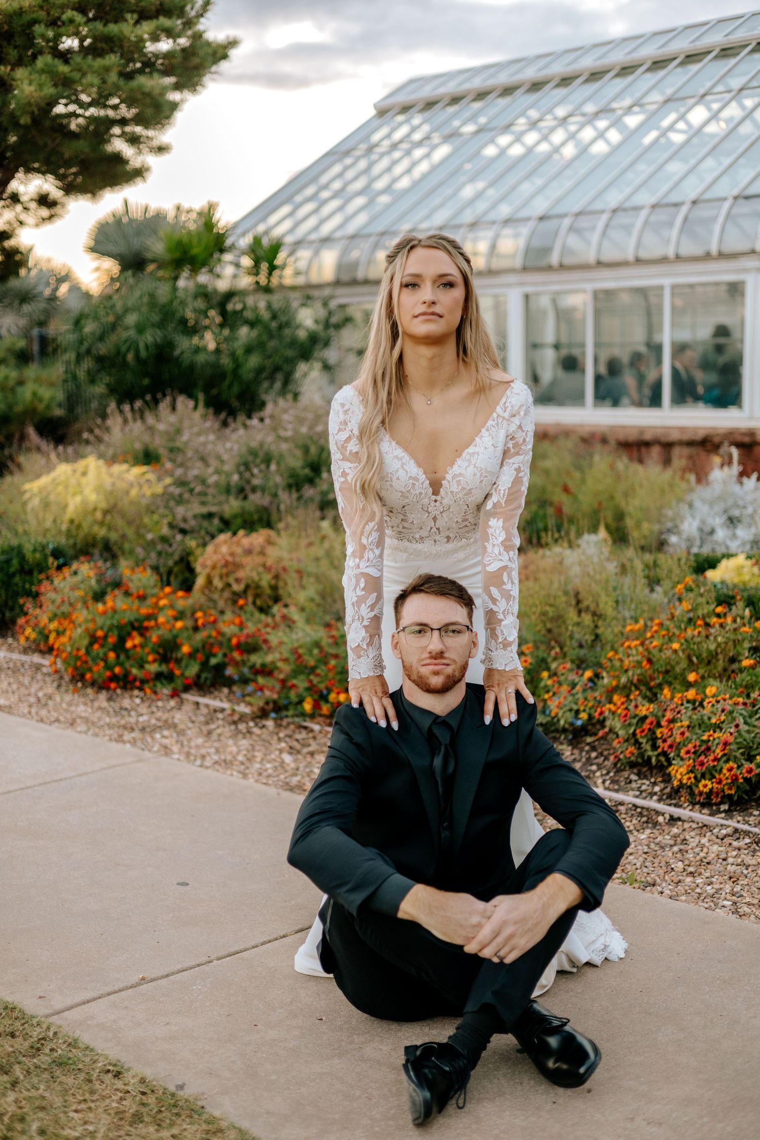 Bride rests hands on seated grooms shoulders in stylish portrait outside botanical greenhouse.