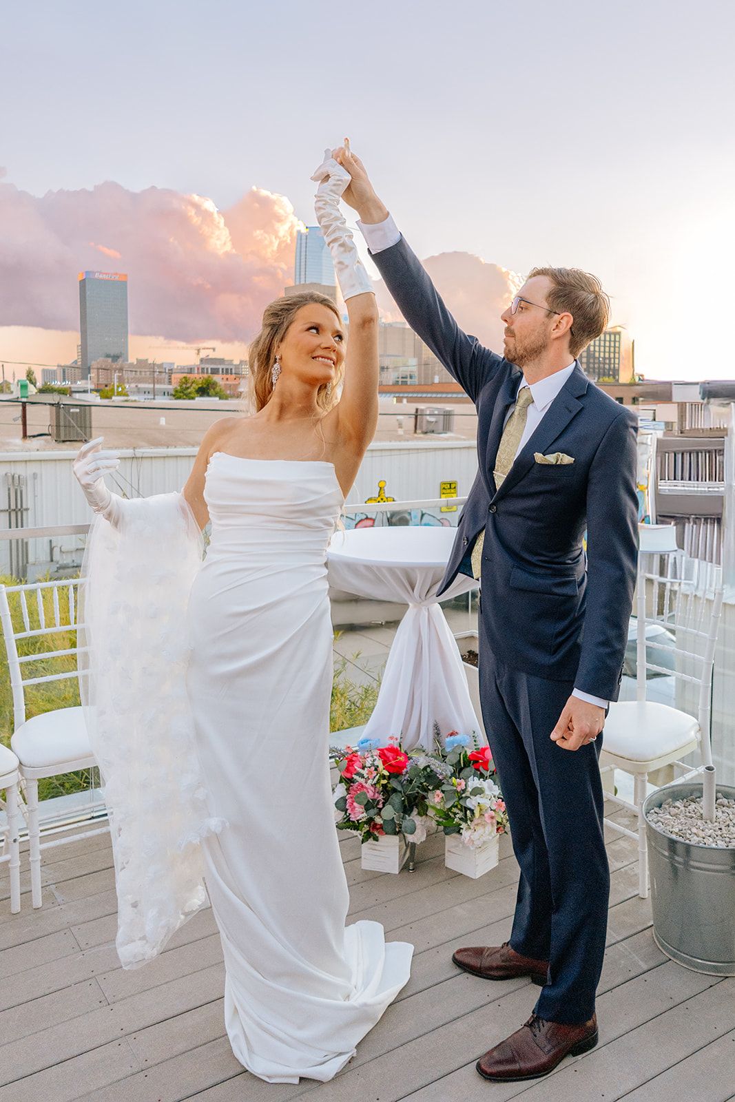 Bride and groom dance together on rooftop terrace during pink sunset with urban skyline in background.