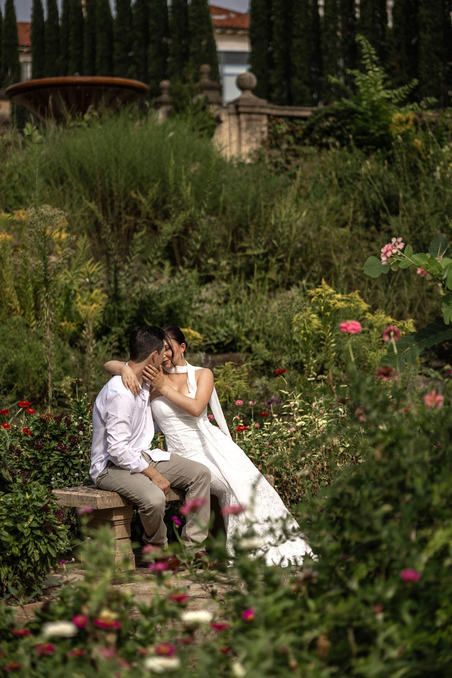 Bride and groom share a romantic kiss on a garden bench surrounded by lush greenery and colorful flowers.