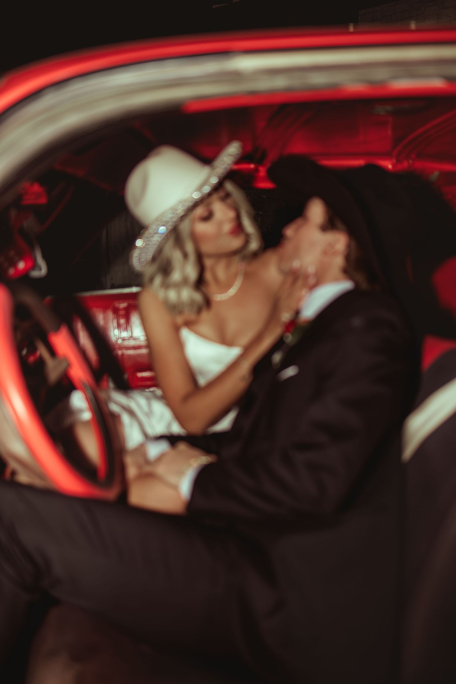 A bride and groom share a romantic kiss inside a vintage red car, wearing cowboy hats on their wedding day.