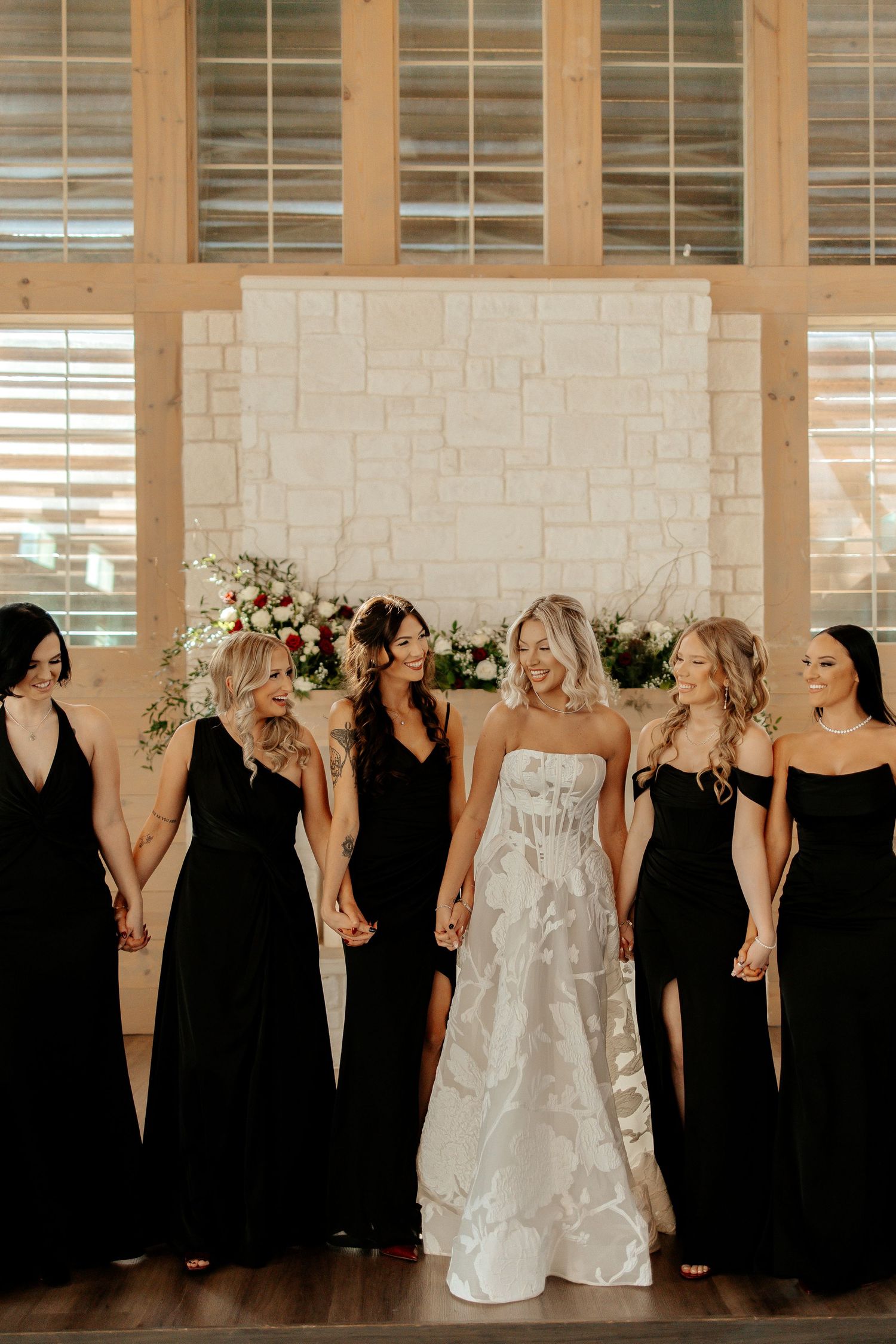 Bride in white floral gown stands with five bridesmaids in black dresses before a stone backdrop.