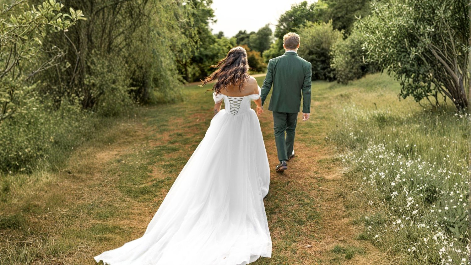 Bride and groom walking hand in hand through a lush green garden path, bride wearing a flowing white off-shoulder gown.