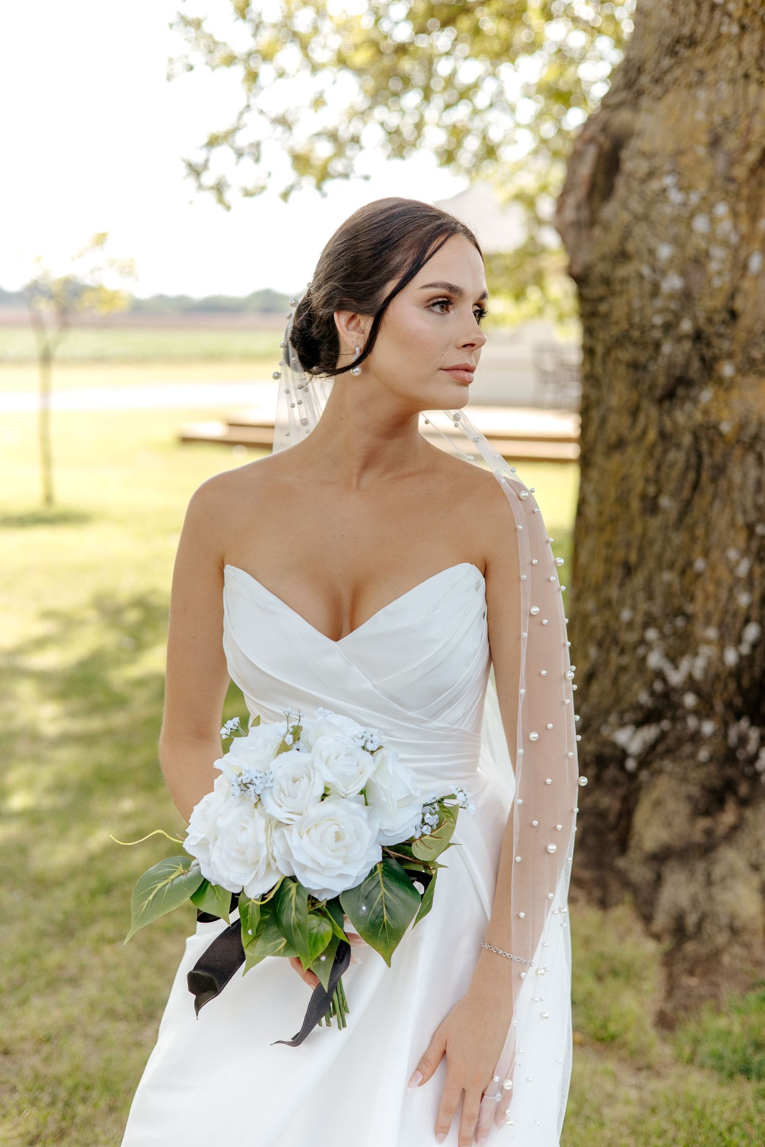 Elegant bride in strapless white dress holding white rose bouquet standing beside a large tree.