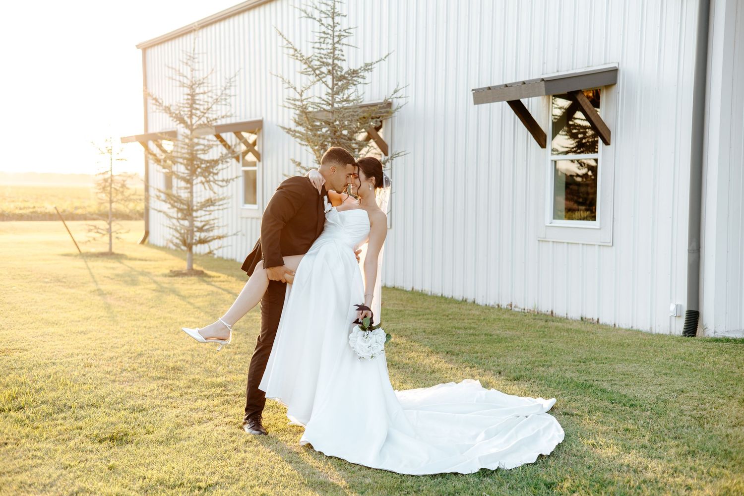 Groom dips bride in white gown outside white barn venue during golden hour wedding portraits.