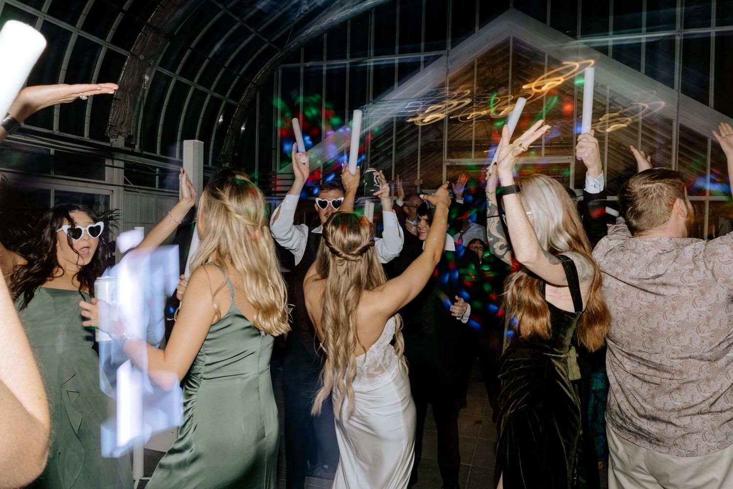 Bride and bridesmaids in green dresses dancing together at a greenhouse wedding reception.