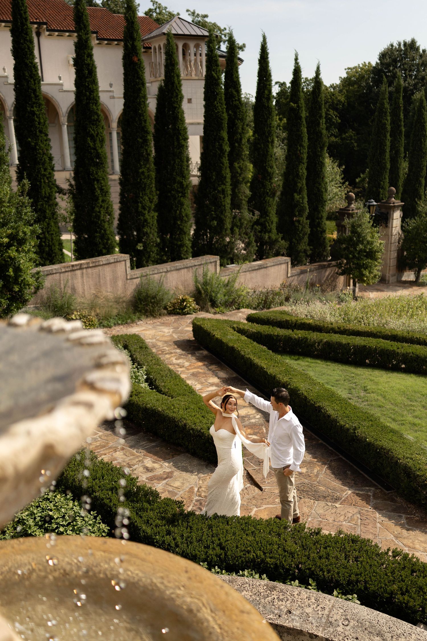 Bride and groom dancing in an elegant formal garden with cypress trees and a historic villa backdrop.