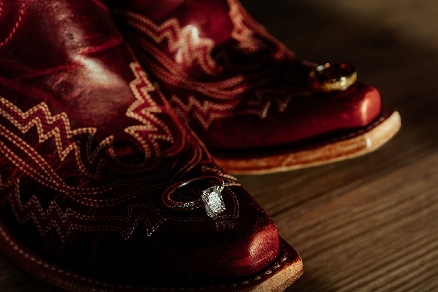 Red cowboy boots with gold stitching displayed with diamond engagement ring and wedding band on wooden floor.