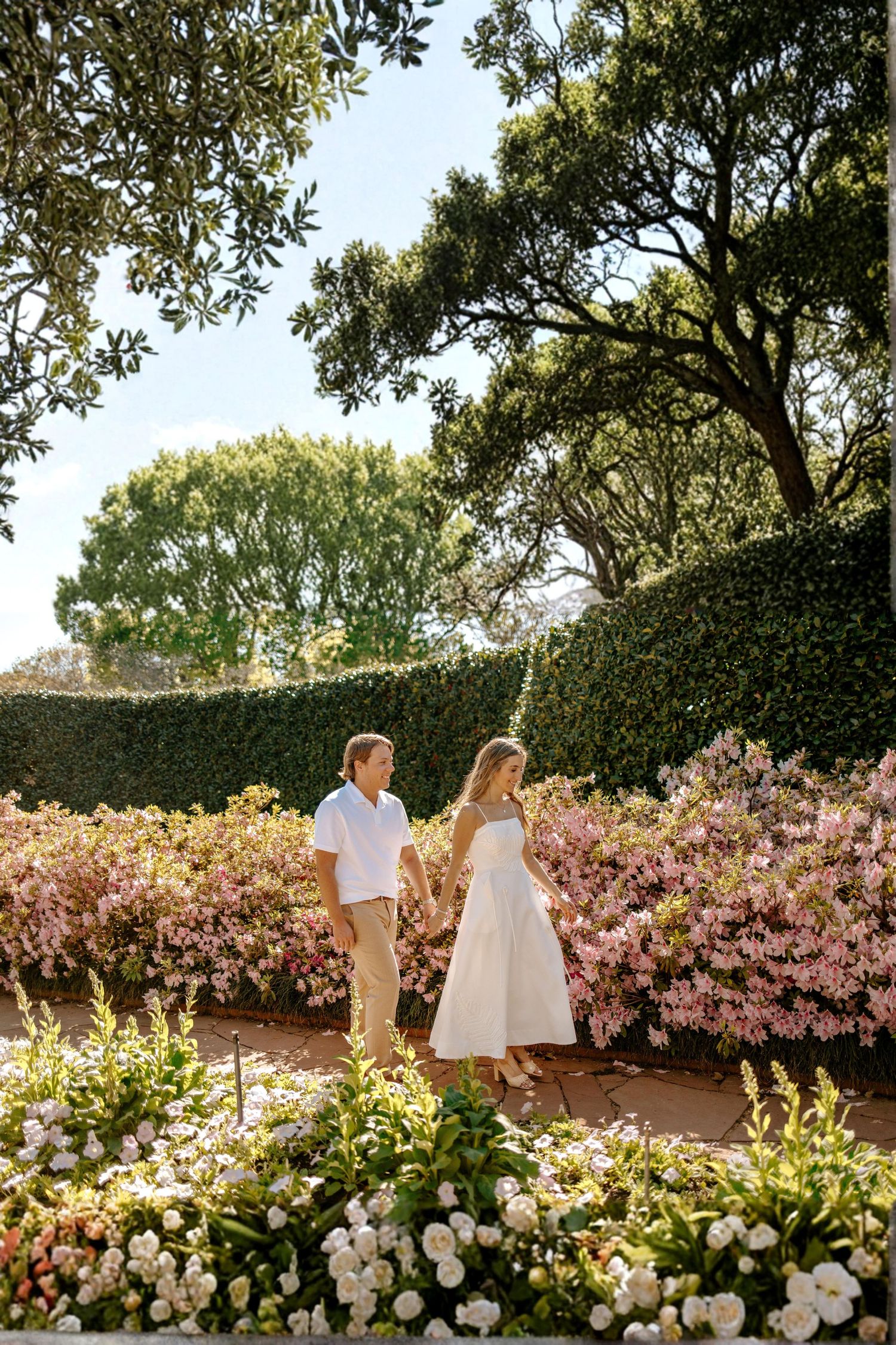 Couple holding hands in a lush garden surrounded by blooming pink flowers and green hedges on a sunny day.