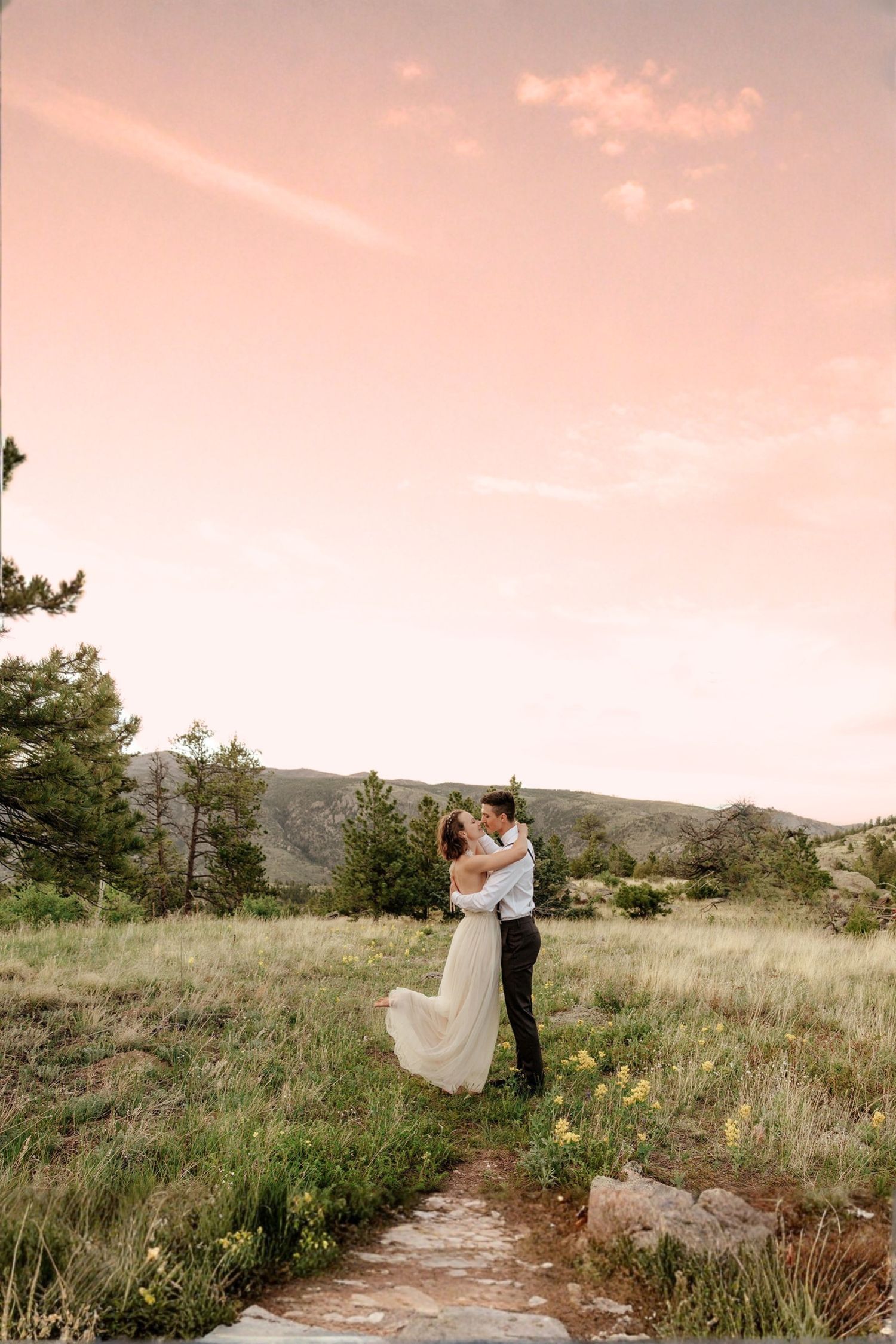 Bride and groom embracing in a mountain meadow at sunset with wildflowers and pink sky.