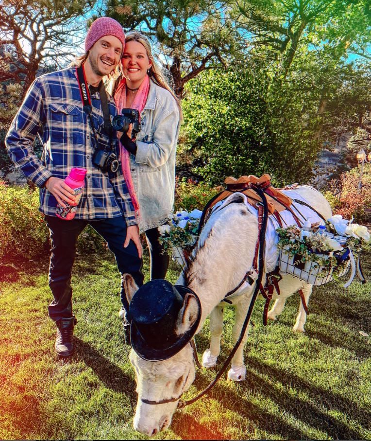 A smiling couple poses outdoors with a decorated white donkey wearing a floral saddle on a sunny day.