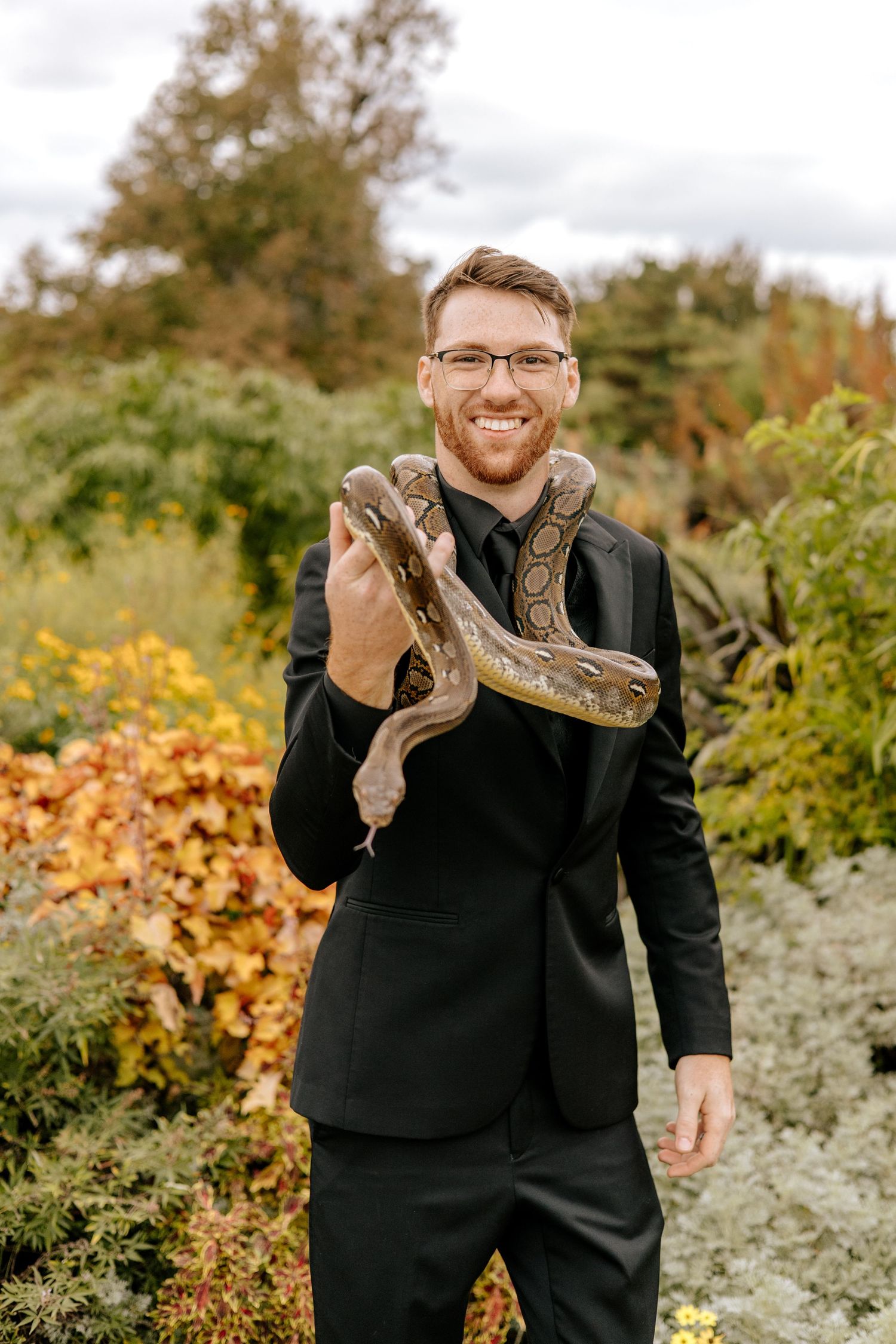 Smiling groom in black suit holds large boa constrictor snake while posing in colorful autumn garden setting.