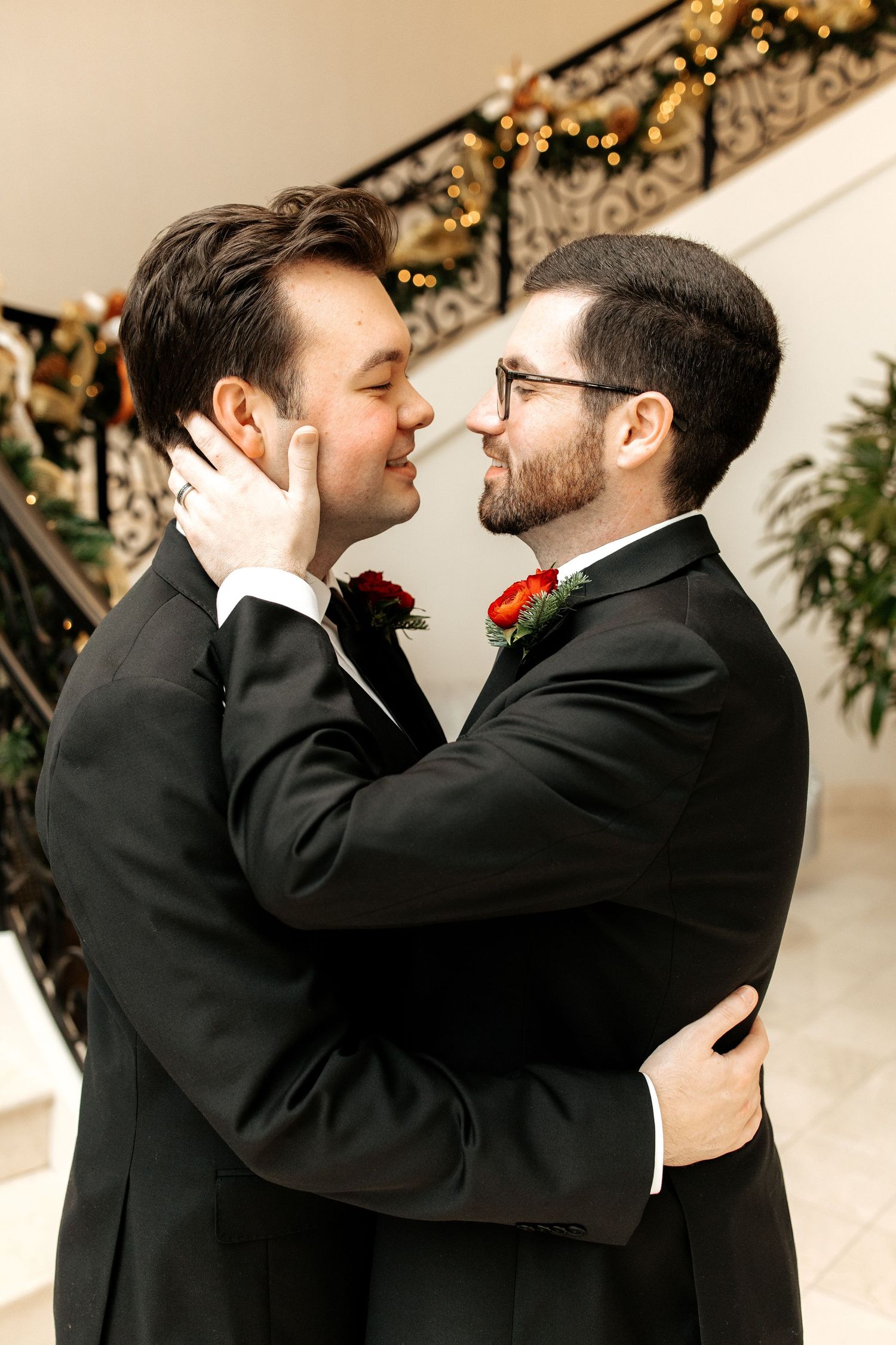 Two grooms in black tuxedos with red boutonnieres share a tender moment on their wedding day.