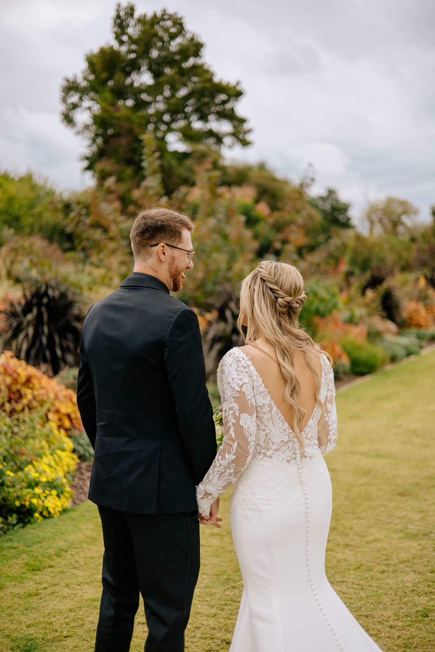 Bride in lace long-sleeve gown and groom in black suit walk hand-in-hand through a colorful autumn garden setting.