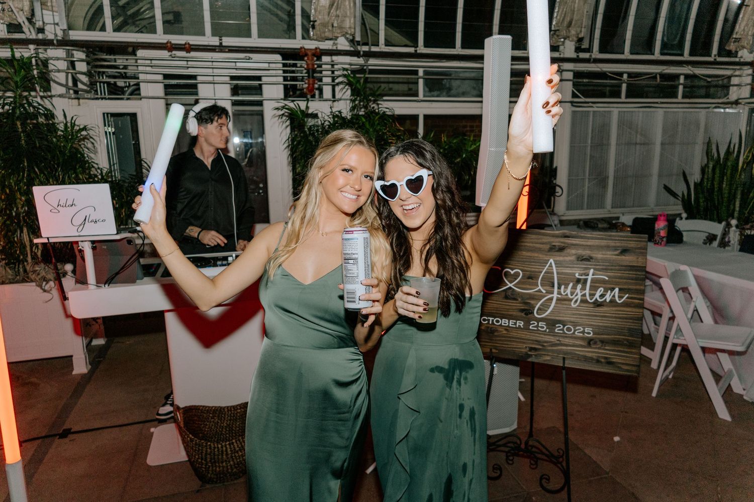 Two bridesmaids in green dresses holding glow sticks and drinks at a wedding reception.