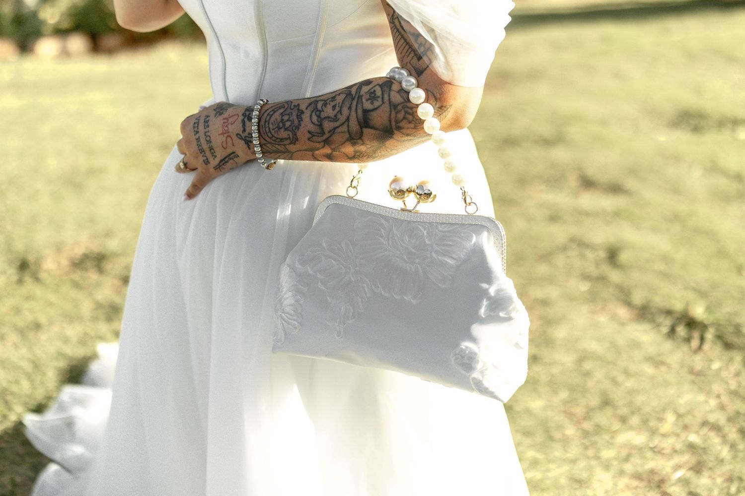 Tattooed bride in white dress holding a silver Louis Vuitton clutch purse outdoors.