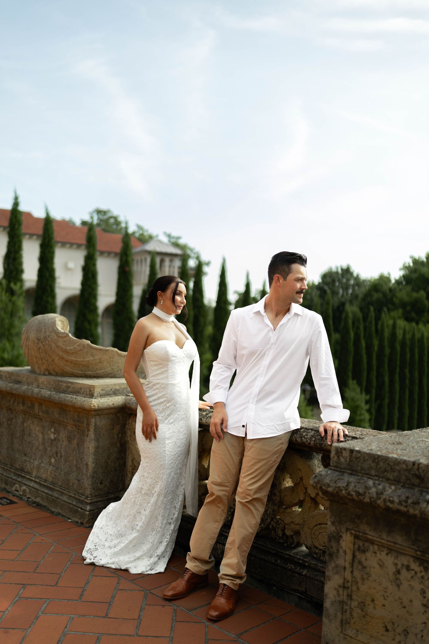 Bride in white lace gown and groom in white shirt pose on a stone terrace with cypress trees.