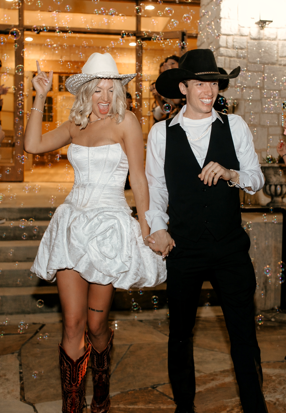 A joyful couple in cowboy hats exits amid bubbles, bride in white mini dress with boots, groom in black vest.