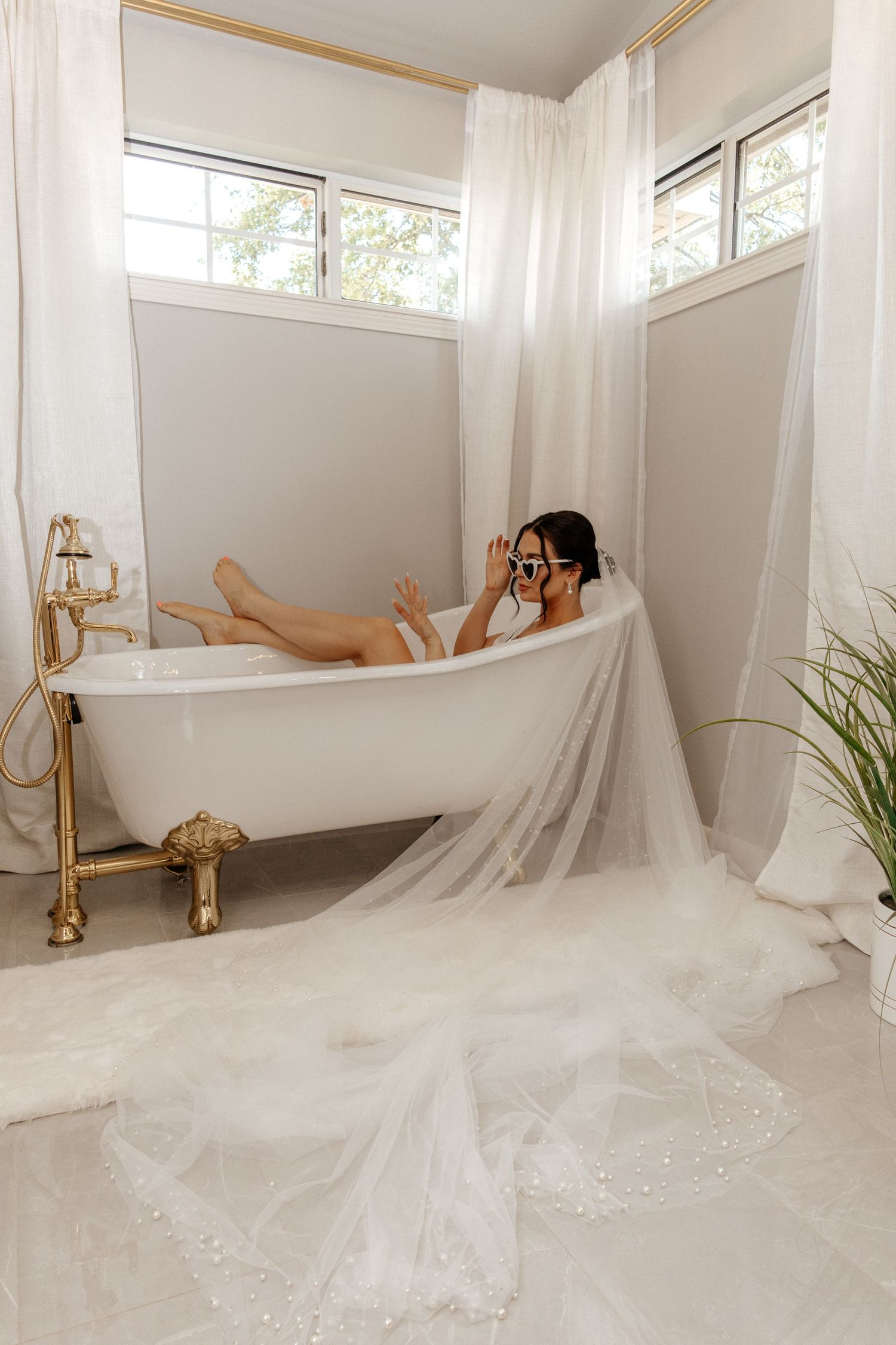Bride in white lingerie and long veil relaxing in a vintage clawfoot bathtub with gold fixtures in bright bathroom.