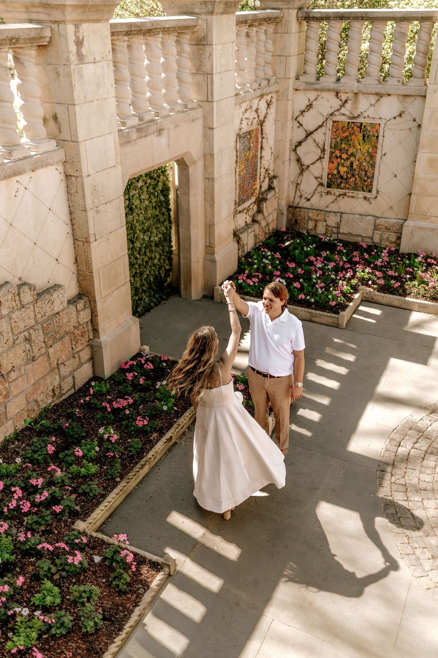 Couple dancing in a sunlit elegant stone courtyard garden with pink flowers and ivy walls.
