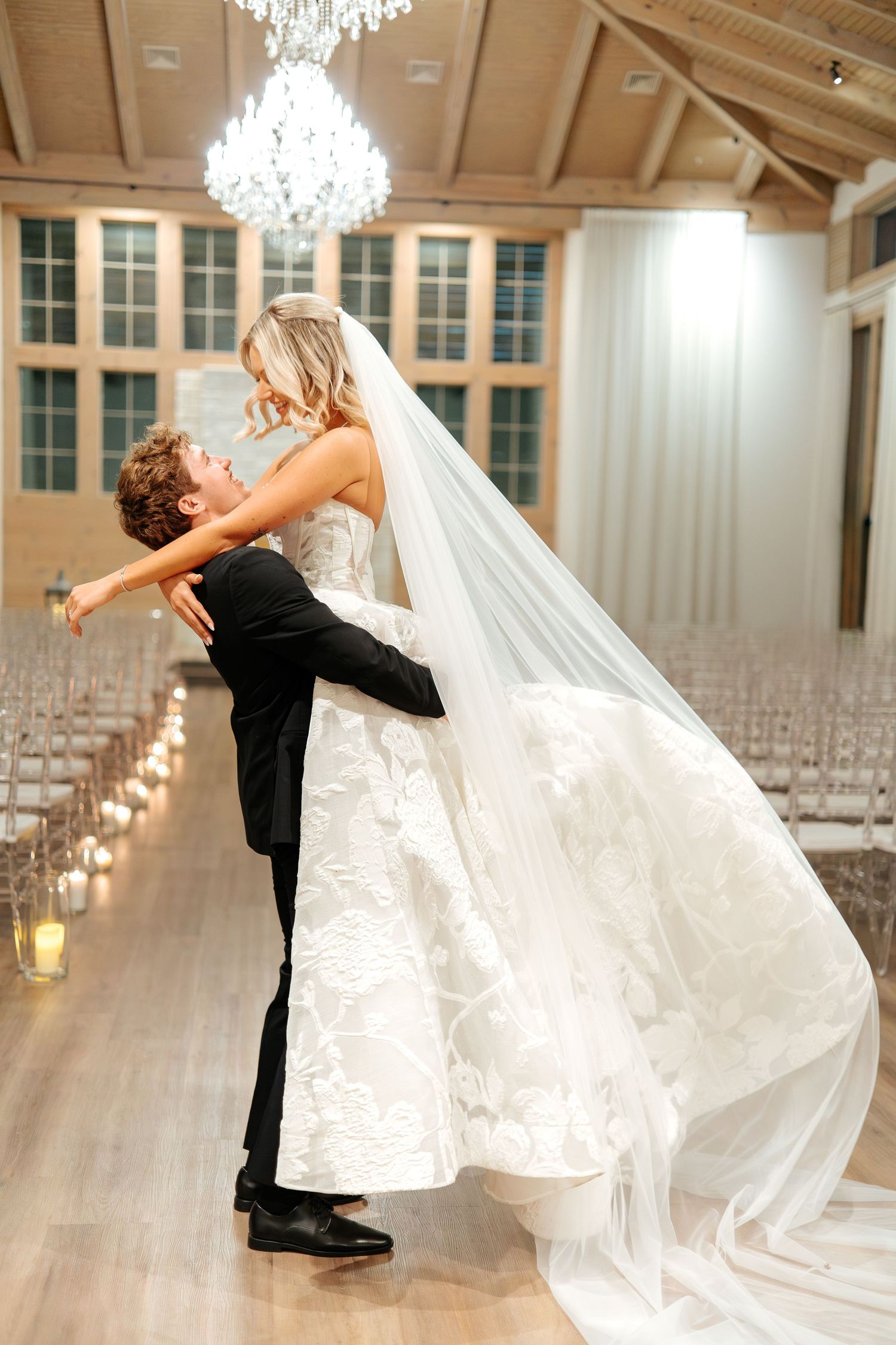 Groom lifts bride in stunning floral ballgown in elegant chandelier-lit wedding ceremony venue.