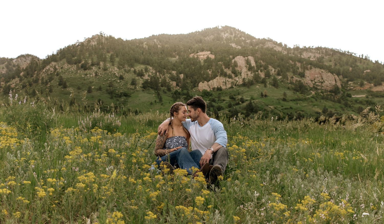 Couple sitting together in a wildflower meadow with mountain backdrop during golden hour engagement session.