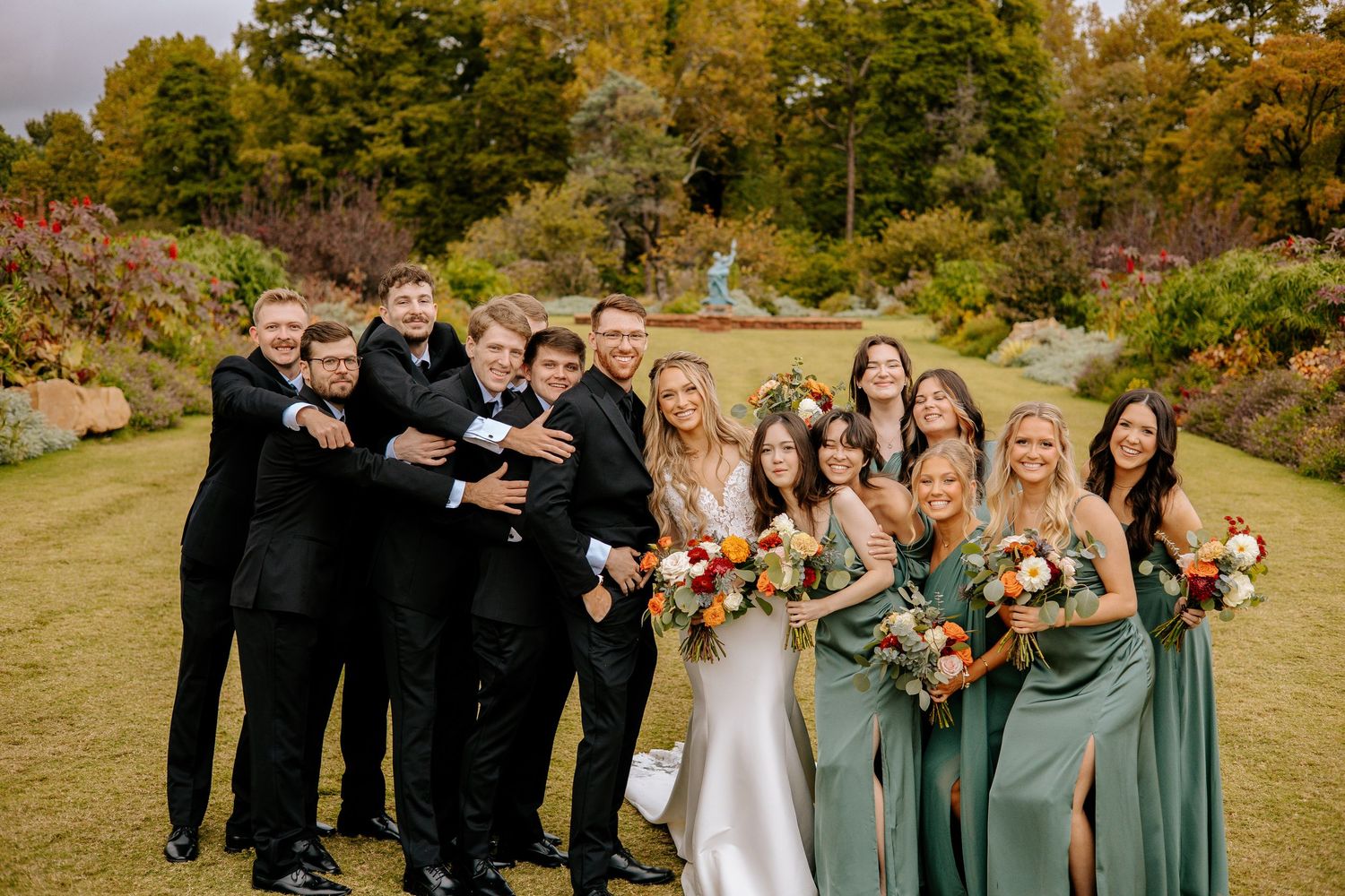 Wedding party poses outdoors in fall garden, groomsmen in black suits and bridesmaids in sage green dresses holding bouquets.