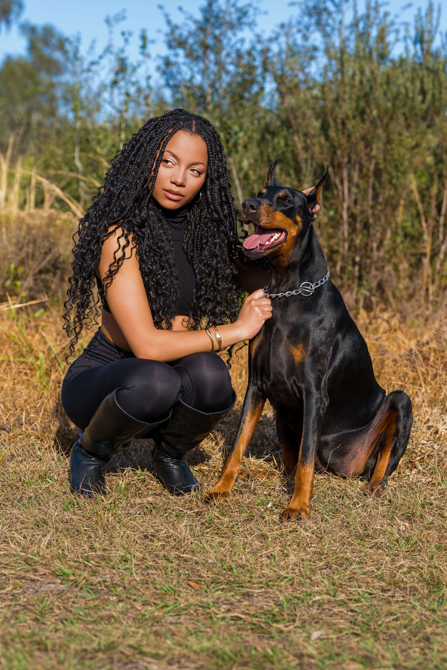 A woman with long braids crouches beside a Doberman dog in a dry grassy field.