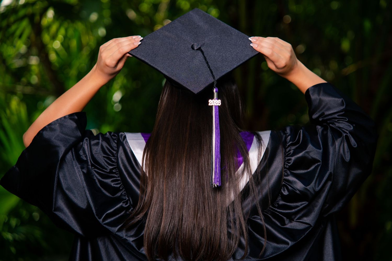 Graduate in black cap and gown holds mortarboard hat, viewed from behind with purple tassel, surrounded by green foliage.