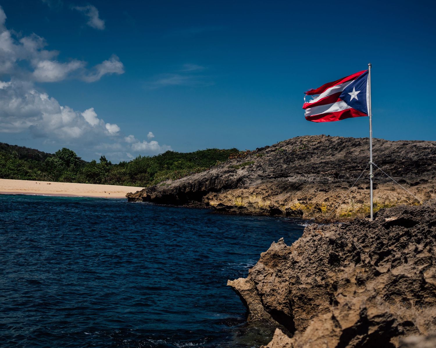 Puerto Rican flag waving on rocky coastline with blue ocean and beach in background.