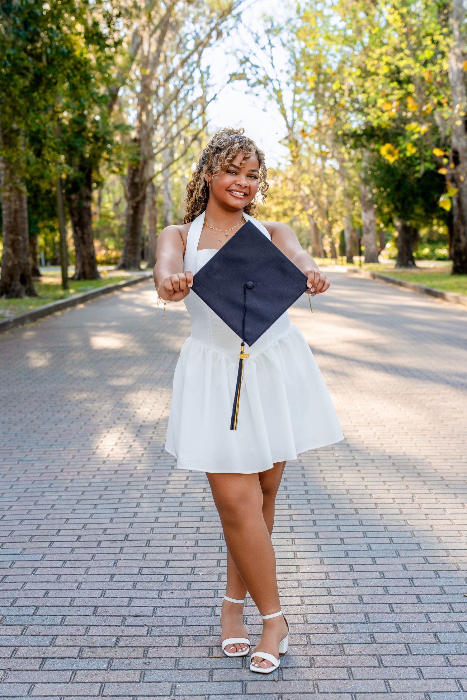 Smiling graduate in white dress holds black mortarboard cap on tree-lined brick pathway.