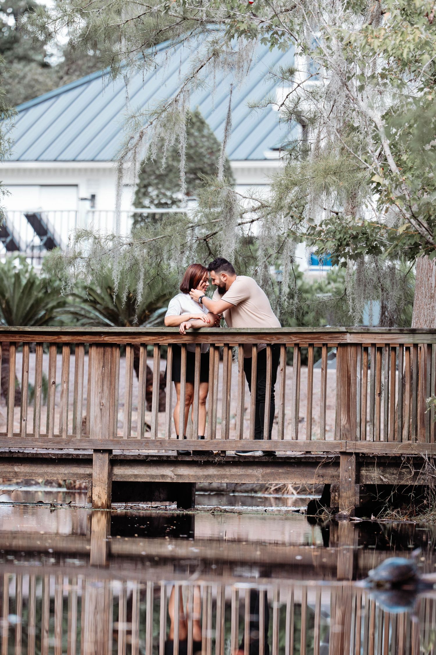 Romantic couple sharing intimate moment on rustic wooden bridge over pond surrounded by lush trees.